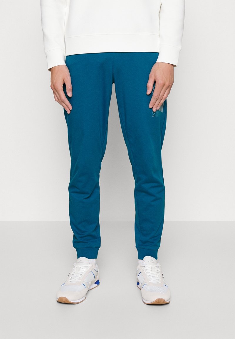 Man står iförd teal joggerbyxor, vita sneakers med blå accenter och en vit långärmad skjorta, mot en enfärgad ljus bakgrund.