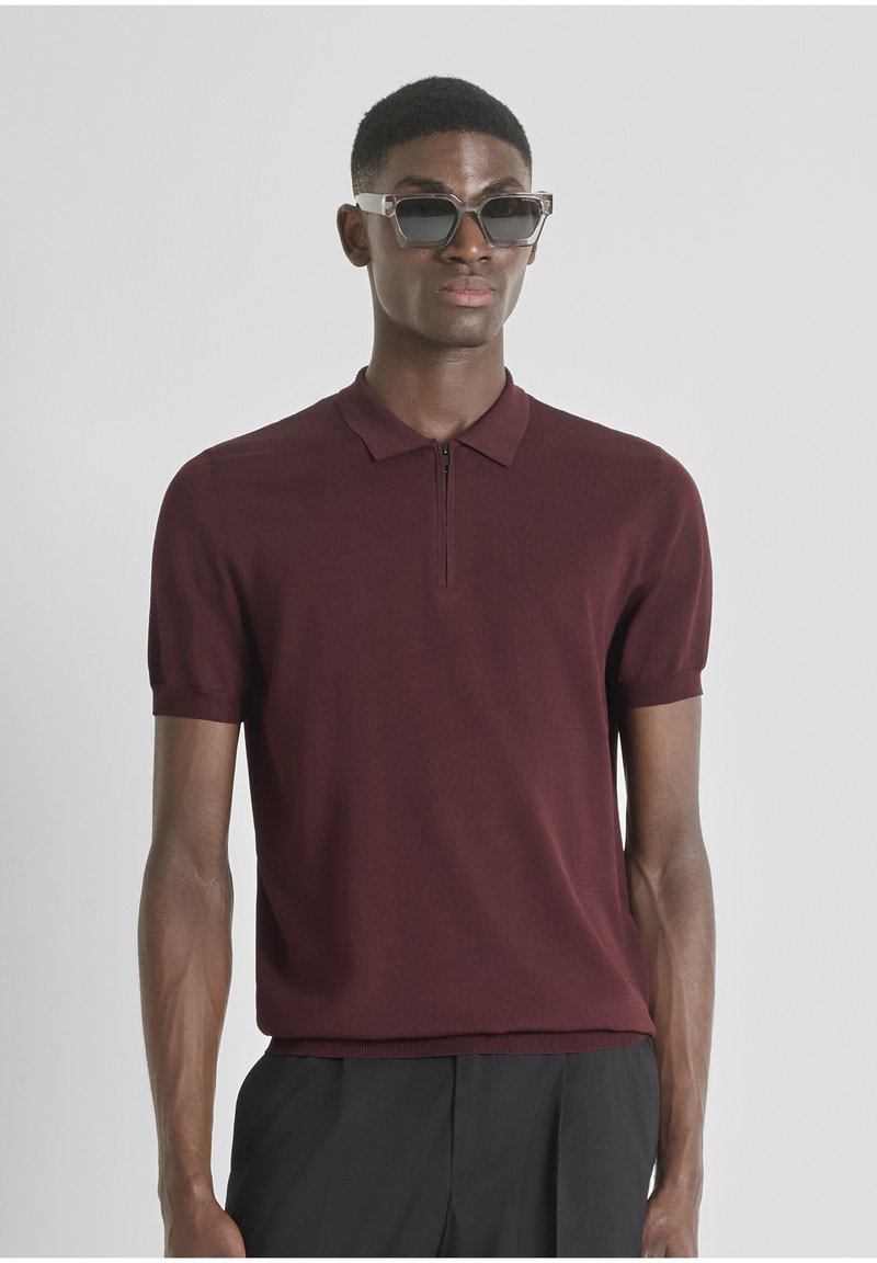 Polo shirt bordeaux à manches courtes avec une fermeture éclair, fabriqué dans un tissu lisse. Porté avec des lunettes de soleil claires et angulaires. Design simple et moderne.