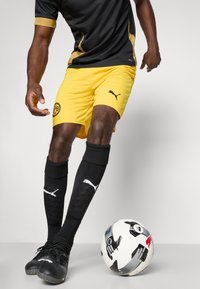 Svart og gul Puma sportsdrakt med mesh-sider, gule shorts med logo, svarte knelengde-strømper, svarte fotballsko og en mønstrete fotball.