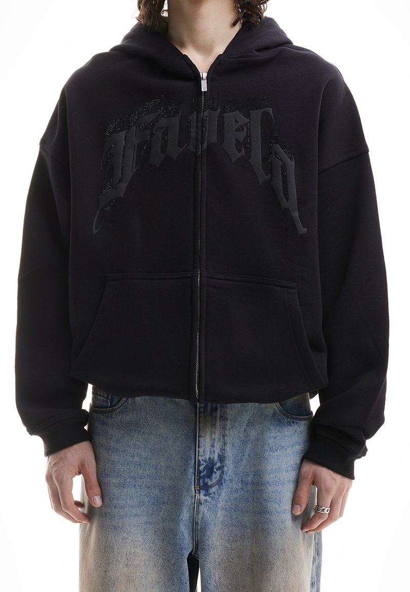 Personne portant un sweat à capuche noir zippé avec le texte "Graveyard" et un jean délavé bleu clair, se tenant les mains relâchées le long du corps.