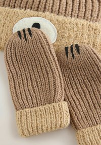 Moufles en tricot marron avec un design texturé, dotées d'un poignet beige et d'accents de point noir sur les doigts, accompagnées d'un bonnet assorti.