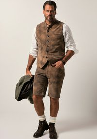 Chaleco de ante marrón con pantalones cortos a juego, camisa blanca debajo y chaqueta verde caída. Botas marrones con cordones y calcetines crema completan el look.