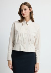 Krótkie, zapinane na guziki w beżowym kolorze, z zaostrzonym kołnierzykiem, długimi rękawami i marszczeniem w talii. Połączone z ciemną spódnicą.