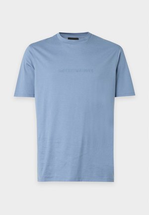 Un jeune homme est debout, portant un t-shirt bleu clair à manches courtes, un jean gris foncé usé, et des baskets en daim marron sur un fond uni.
