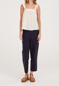 Jasna kremowa bluzka bez rękawów z falbankowymi ramiączkami, zestawiona z dopasowanymi ciemnoniebieskimi spodniami i beżowymi sandałami z grubą podeszwą.