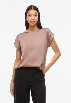 Donna con capelli scuri lisci indossa una blusa lilla a maniche corte e larghe e pantaloni neri a vita alta, in piedi con una mano in tasca.