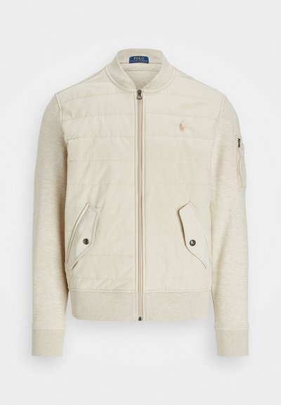 Casaco bomber bege claro, frente acolchoada com mangas lisas em creme, dois bolsos frontais com aba, bolso com fecho no braço, punhos canelados.