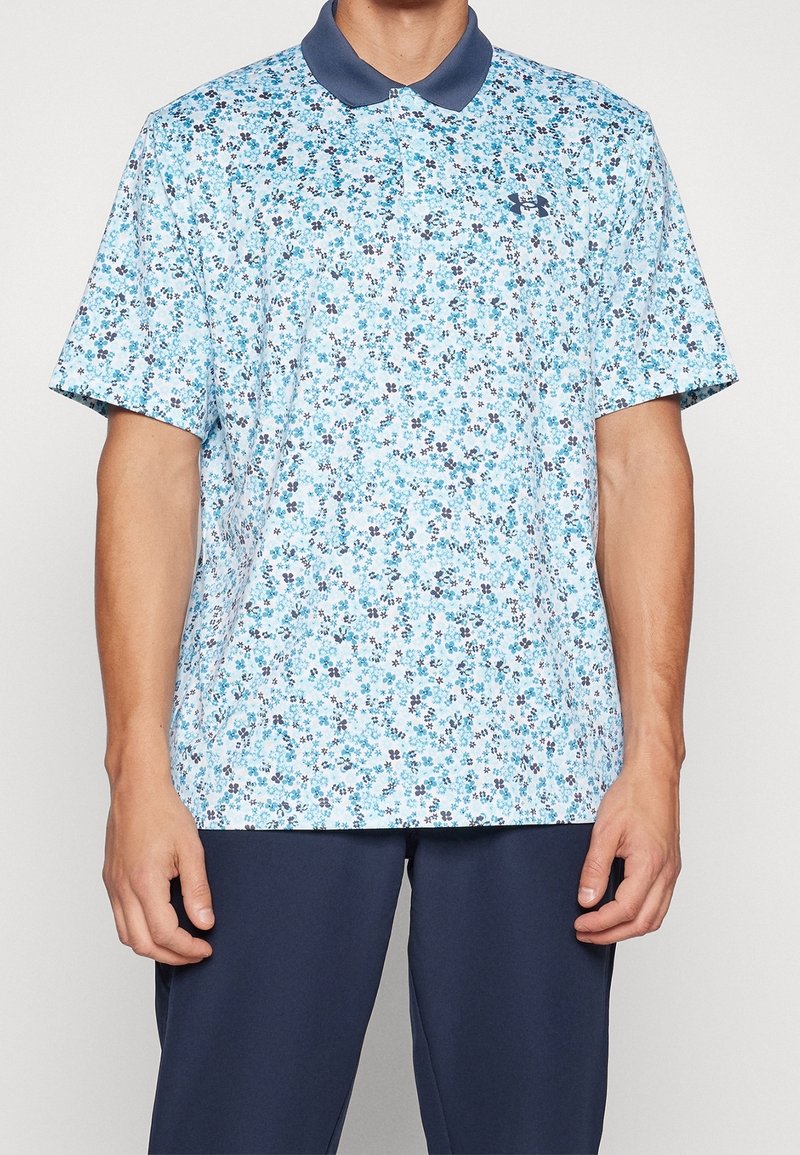 Homme portant un polo à manches courtes bleu clair à motifs floraux avec un col bleu marine et un pantalon bleu marine, debout devant un fond uni.