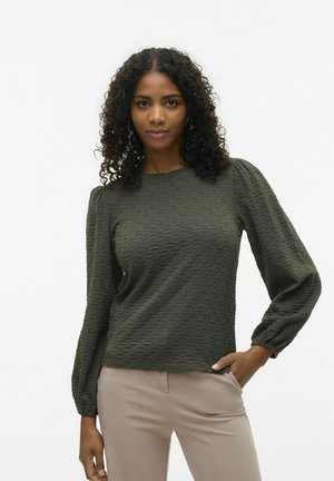 Mörkgrön texturerad topp med långa puffärmar, rund halsringning och en något oversized passform. Kombinerad med beiga byxor.