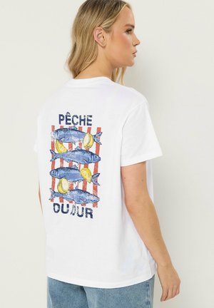 Femme blonde portant un t-shirt blanc avec un motif de poisson bleu et citron sur des rayures rouges et le texte français "Pêche du jour" au dos.