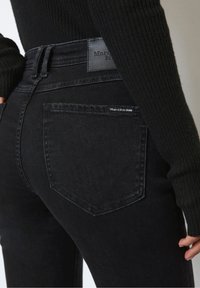 Jean en denim noir taille haute, avec une étiquette en cuir sur la ceinture arrière et une petite étiquette de marque au-dessus de la poche.