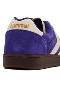 Fioletowy zamszowy sneaker z kremowym obcasem i złotym logo. Posiada teksturowaną brązową podeszwę i białe paski akcentujące.