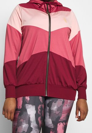 Femme portant une veste zippée bicolore bordeaux et rose avec des leggings à motifs dans des tons de rose, noir et gris.