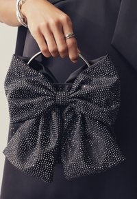 Juoda satino rankinė su dideliu kalnų krištolu puoštu lanku, sidabrine rankena ir tekstūruotu dizainu. Rankinė turi minkštą, blizgią apdailą.