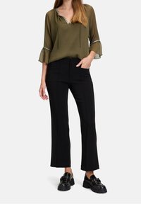 Blouse légère vert olive avec encolure en V, manches évasées et bordure argentée, associée à un pantalon noir ajusté et des chaussures noires épaisses.