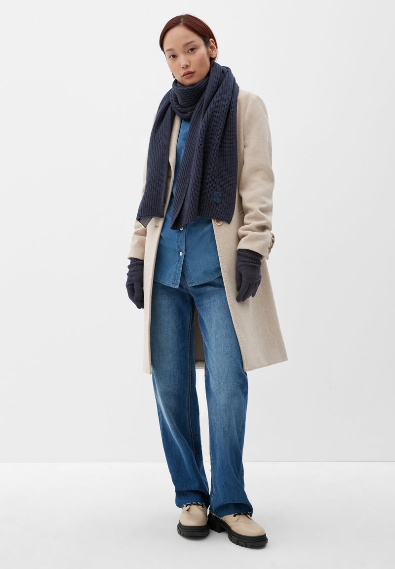 Manteau en laine beige, chemise en denim bleu, écharpe en tricot bleu foncé, gants navy, jean bleu à jambes larges, chaussures beiges avec semelles noires, détail de marque minimal.