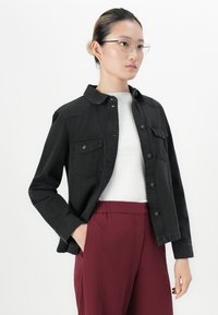 Ungdom med briller, iført en svart knapp-up jakke over en hvit topp og vinrøde bukser, står med en hånd i lommen mot hvit bakgrunn.