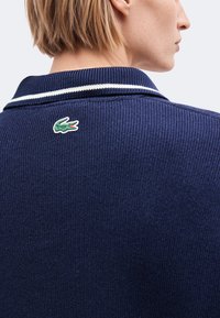 Cheveux blonds courts et profil de côté portant un pull en maille bleu marine avec une bande blanche sur le col et un petit logo crocodile vert dans le dos.