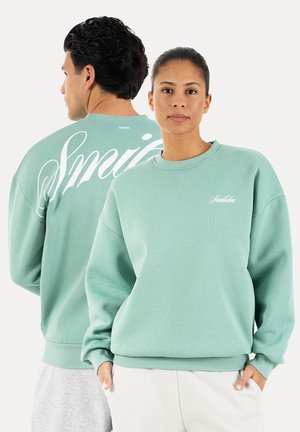 Mężczyzna i kobieta w pasujących jasnoliściach bluzach z białym logo pisanym kursywą, kobieta zwrócona przodem, mężczyzna tyłem prezentujący duży wzór na plecach.