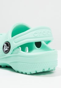 Turquoise Crocs met een gestructureerd oppervlak, achterband en rond zwart logo-detail. Lichtgewicht materiaal, met afvoergaten en een gevormde pasvorm.