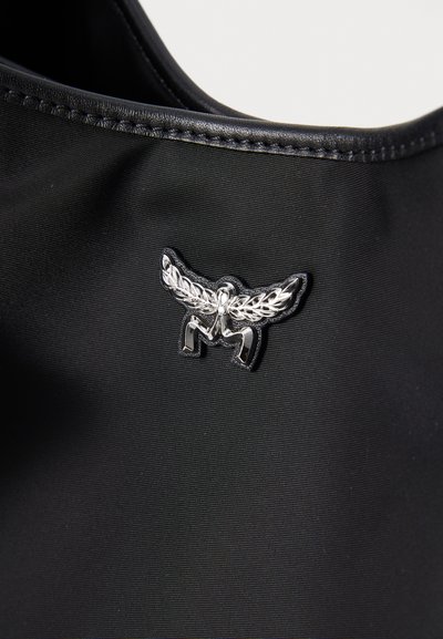 Musta kangas, jossa on sileä pinta, esittelee hopeista emblemia siipineen ja 'M'-yksityiskohdalla, kiinnitettynä sivulle.