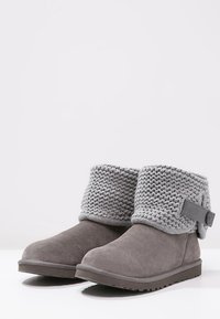 Par de botas de tobillo de ante gris con puños de punto doblados y detalle de correa con botón sobre un fondo blanco.