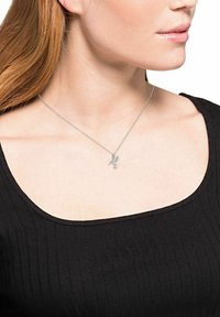 Collar de tono plateado con un colgante de pájaro, colgado de una fina cadena. Lleva puesto sobre una camiseta negra de canalé, resaltando el diseño del collar.