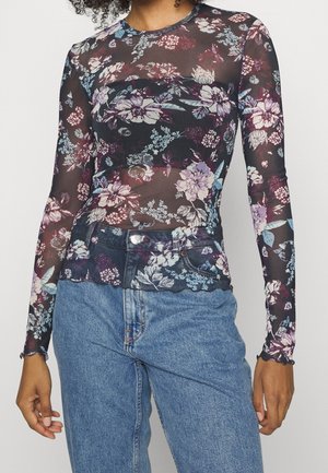 Femme portant un haut noir transparent à manches longues avec des motifs floraux roses et violets, associé à un jean bleu taille haute.