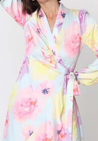 Femme portant une robe portefeuille pastel avec de grands motifs floraux roses, jaunes et violets, nouée à la taille avec des manches longues.