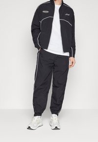 Zwarte trainingsjas met witte pipingdetails, gecombineerd met bijpassende broek. Model draagt een wit t-shirt eronder en witte sportschoenen.