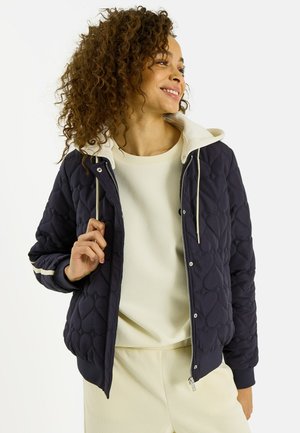 Femme aux cheveux bouclés portant une veste matelassée bleu marine sur un sweat à capuche crème et un pantalon assorti, souriant et regardant sur le côté.