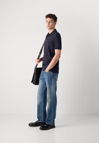 Jūrinės polo marškiniai, šviesiai mėlyni džinsai ir juodi batai. Juodas besiūlis krepšys su dirželiu. Modelis stovi prieš šviesiai pilką foną.