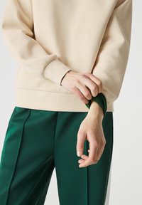 Sudadera beige con un acabado texturizado, con un dobladillo ajustado. Combinada con pantalones verdes que presentan pliegues y una textura suave.