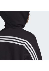 Personne portant un sweat à capuche noir avec trois bandes blanches traversant le dos et l'épaule.