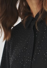Blusa nera in tessuto testurizzato con piccoli accenti luccicanti, colletto classico e chiusura a bottone. Materiale morbido e leggero.