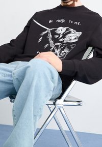 Sudadera negra con un gráfico de un perro con una correa, con letras blancas que dicen "Sé amable conmigo" sobre el diseño. Jeans azul claro.