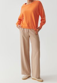 Osoba ubrana w pomarańczową bluzkę z długim rękawem i sznurkiem w talii, beżowe spodnie o szerokich nogawkach oraz beżowe trampki, stojąca z rękami w kieszeniach.