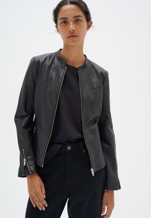 Chaqueta de cuero negra con un escote redondo, cierre frontal y puños con cremallera; llevada sobre una camiseta oscura y combinada con pantalones negros.