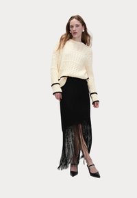 Maglione crema a trama cable con rifiniture nere, abbinato a una gonna nera con dettagli in frange e tacchi neri a punta. La modella posa in posizione centrale.
