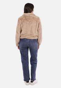 Giacca in pelliccia sintetica beige con colletto, indossata sopra jeans di denim blu e sneaker bianche. La giacca presenta una superficie morbida e rigata.