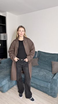 Rudas ilgas paltas, juodi marškinėliai ir juodos flared džinsai; stovi ant šviesios medienos grindų šalia mėlyno apmušto sofos su raštuotais pagalvėlėmis.