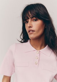 Lichtroze poloshirt met kraag, korte mouwen, twee voorzakken en gouden knopenaccenten op de sluiting. Glad, gestructureerd weefsel.
