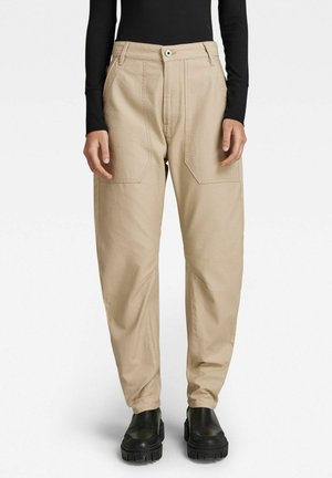 Personne portant un pantalon beige taille haute avec de grandes poches avant, un haut noir à manches longues côtelé, et des bottes noires à semelle épaisse, debout.