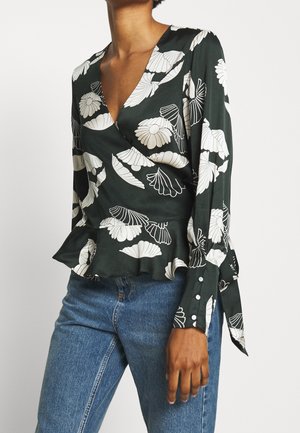 Donna che indossa una camicetta avvolgente verde scuro con stampa floreale bianca, orlo a peplo, maniche lunghe con polsini con bottoni, abbinata a jeans blu.