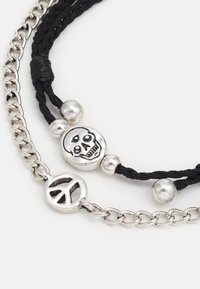Pulseira de corda preta trançada com contas prateadas, um encanto de caveira e um símbolo da paz em metal. Materiais texturizados e contrastantes.