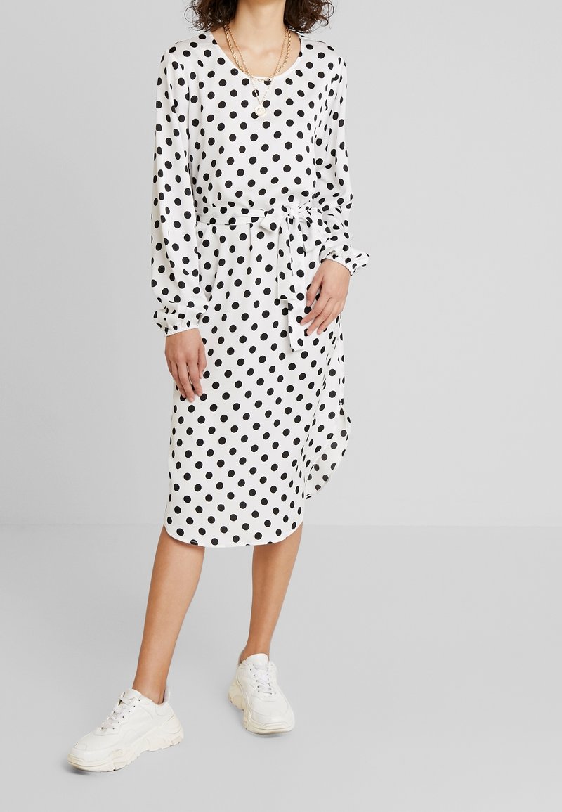 Ein gepunktetes Kleid in Weiß und Schwarz, mit langen Ärmeln, einem taillierten Design und einem knielangen Saum. Kombiniert mit klobigen weißen Sneakers.