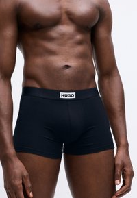 Fit, dark-skinned male torso wearing black boxer briefs with a white waistband label reading "HUGO" against a plain white background.