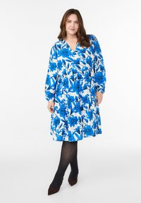 Sinine lilleline mähkiv kleit pikkade varrukatega, V-kaelusega, elastse vöökoha ja laieneva seelikuosa ning mustade sukkpükste ja pruunide kontsakingadega.