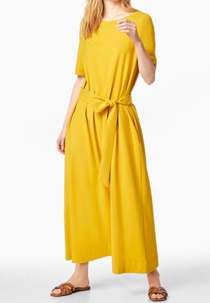 Femme portant une robe midi jaune moutarde à manches courtes avec une ceinture nouée à la taille, associée à des sandales plates marron sur fond blanc.