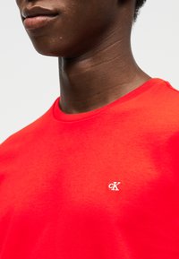 Κοντινό πλάνο ατόμου που φοράει φωτεινό κόκκινο t-shirt με μικρό λευκό λογότυπο "cK" στο αριστερό στήθος πάνω σε απλό ανοιχτό φόντο.